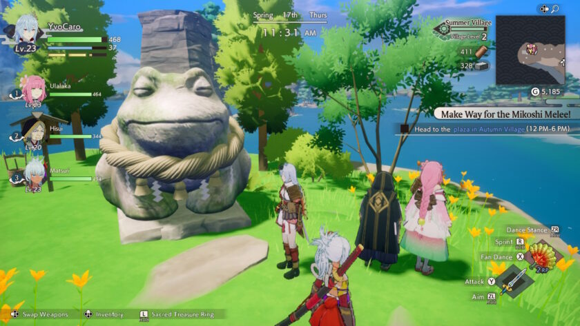 A serene moment in Rune Factory: Guardians of Azuma where the party gathers beside a large frog statue wrapped in sacred rope, overlooking a lake. Yellow flowers dot the grass and trees sway under a bright blue sky. A quest prompt titled “Make Way for the Mikoshi Melee!” is visible. Published on: LadiesGamers