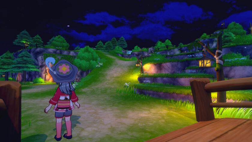 A screenshot from Story of Seasons: Grand Bazaar shows the female protagonist standing on a dirt path at night, gazing up a gently winding hill illuminated by lanterns. She wears a wide-brimmed hat with a flower design, a red jacket, and striped socks. The path is flanked by grassy terraces, wooden fences, and glowing lights, with pine trees and rocky cliffs in the background. The deep blue night sky is dotted with stars and scattered clouds, creating a peaceful and magical atmosphere. Published on: LadiesGamers.